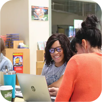 Two students are sitting at a table, smiling and collaborating on a laptop. One student has curly dark hair and glasses, wearing a gray t-shirt, while the other has long dark hair pulled back, dressed in an orange sweater. A teal water bottle and a coffee cup with the words "GET SIPPING" are on the table. In the background, there is a colorful bulletin board and shelves filled with books.
