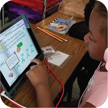 A young girl with curly hair, wearing headphones, sits at a wooden table engaged with a tablet. The screen displays a colorful, interactive educational game with the text: “Catch the Iguana! I'm going to school! Click on the iguana with the word.” Below, there are cartoon iguanas and a hand pointing to one of them. Pencils and a small notebook are visible on the table, along with a stack of books in the background.