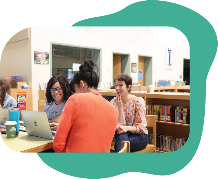 A group of four people are sitting at a table in a library, engaged in a friendly conversation while looking at a laptop. One person wears glasses and has curly hair, another is in a bright orange sweater, and a third is smiling with long hair. Behind them, there are bookshelves filled with colorful books, and the space is bright and welcoming.