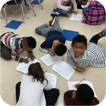 A group of five children sits on the floor of a classroom, focused on their work. They are writing on sheets of paper with pencils. One child with dark curly hair and a striped shirt playfully sticks out their tongue at the camera. The floor has light tiles with blue square sections, and some children are wearing casual clothing like t-shirts and sweatshirts.