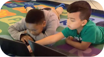 Two children are lying on a colorful rug with letters and numbers, working together on laptops. One child, wearing a gray sweater, rests his head on his arms while looking at the screen. The other child, wearing a green t-shirt, reaches out to touch the laptop. They seem focused and engaged in their activity.
